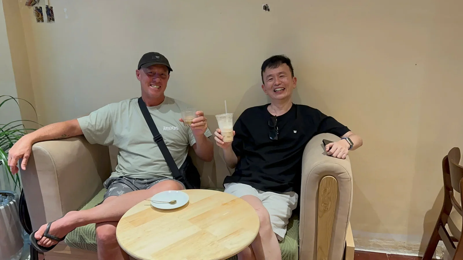 Two guests on the café's beige sofa, raising an iced coffee and an iced tea towards the camera.