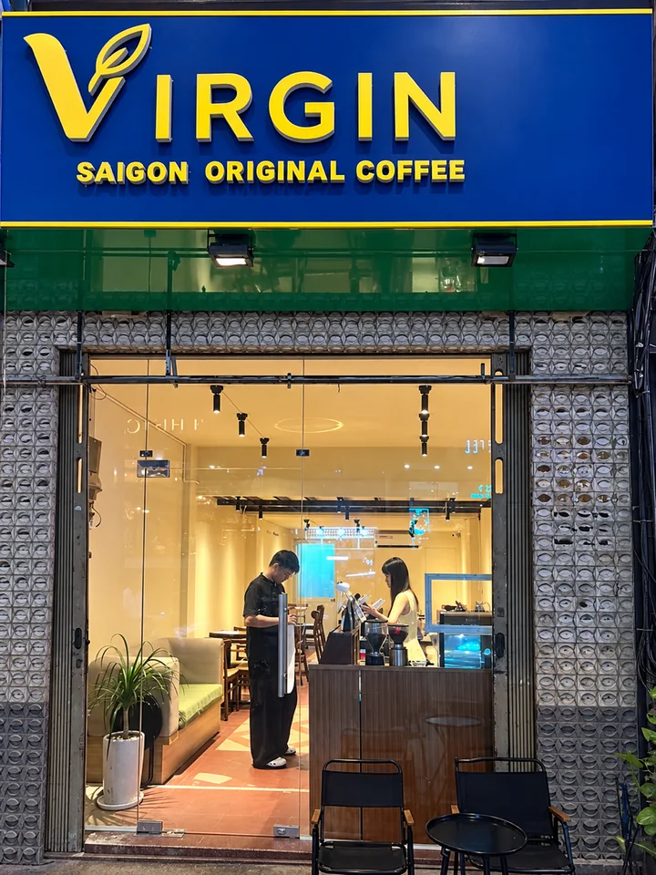 The Virgin Saigon shopfront on Nguyễn Thái Bình after dark. The lit sign reads VIRGIN, Saigon Original Coffee, and two staff are working in the café behind the open front.