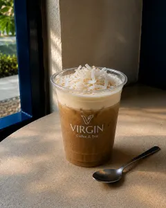 A Virgin Coffee cup of cà phê cốt dừa on a stone table by a blue window frame, pale coconut cream sitting above iced coffee under a heap of shredded coconut, a teaspoon alongside.