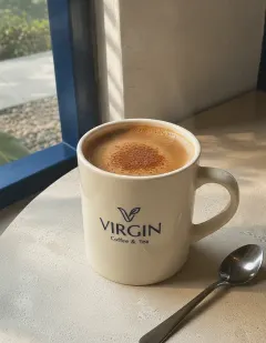 Hot cocoa in a cream ceramic mug with the Virgin logo, golden-tan cocoa dusted with cinnamon, steam rising, a spoon beside it.