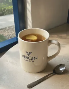 Hot ginger tea in a cream ceramic mug with the Virgin logo, clear amber tea with ginger slivers, steam rising, a spoon beside it.