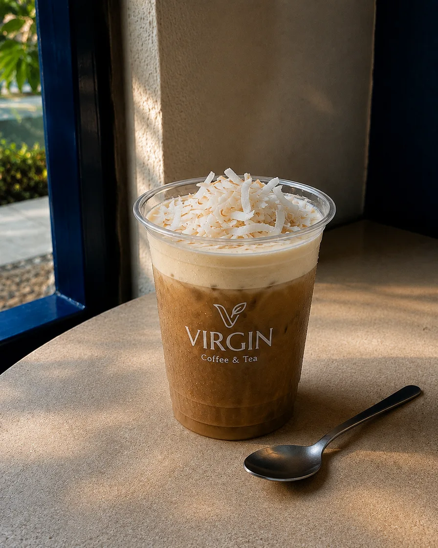 A Virgin Coffee cup of cà phê cốt dừa on a stone table by a blue window frame, pale coconut cream sitting above iced coffee under a heap of shredded coconut, a teaspoon alongside.